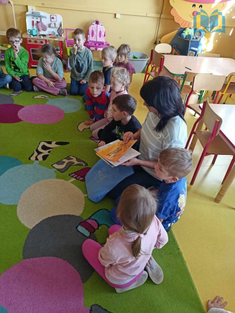 Zdjęcie: Dzieci w przedszkolu siedzą na kolorowym dywanie. Słuchają bibliotekarki, która opowiada im o bibliotece i zasadach wypożyczania książek.